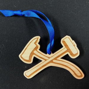 May include: A wooden ornament shaped like two crossed axes, one with a hammer head and the other with an axe head. The ornament is attached to a blue ribbon.