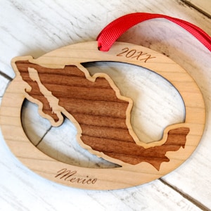 May include: A wooden ornament shaped like the outline of Mexico with the year 20XX engraved on the top and the word "Mexico" engraved on the bottom. The ornament is attached to a red ribbon.