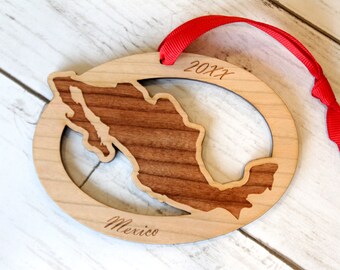 May include: A wooden ornament shaped like the outline of Mexico with the year 20XX engraved on the top and the word "Mexico" engraved on the bottom. The ornament is attached to a red ribbon.