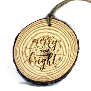 May include: A round wooden ornament with a natural wood grain finish. The ornament has the text "merry and bright" engraved on it in a cursive font. The ornament is decorated with small stars.