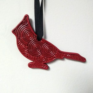 May include: Red cardinal bird ornament with a swirling pattern. The ornament is made of clay and has a black ribbon for hanging.
