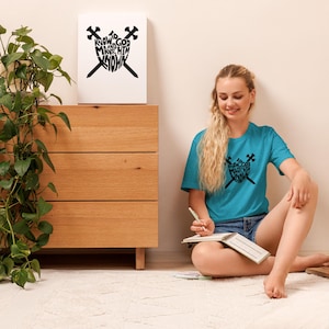 以下が含まれることがあります： 「KNOW TO GOD MAKE HIM KNOW」の文字が入った黒いグラフィックデザインのターコイズ色のTシャツ。デザインは、交差した剣を持つハートの形をしています。白いラグの上に座っている人が着ています。