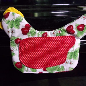 May include: A chicken-shaped fabric oven mitt featuring a white background with a green strawberry plant and red strawberry print. The center of the mitt has a red fabric patch with a white polka dot pattern, and a yellow fabric comb on top.