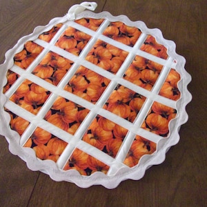 May include: White fabric hot pad with a scalloped edge. The pad is decorated with a grid pattern of orange and brown pumpkin fabric.