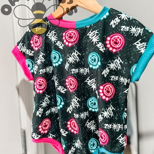 May include: Black and white baby onesie with a pink and blue trim. The onesie has a repeated pattern of the band Blink-182's logo and the words "Blink-182" and "28-NITA".