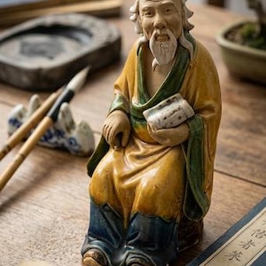 May include: A ceramic Shiwan Mudman figurine depicting an elderly person with a long white beard, wearing a yellow robe with green trim and blue trousers, holding a small white scroll, accompanied by a tag labeled SHIWAN MUDMAN Hong Kong.