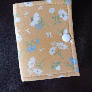 May include: A small yellow fabric pouch with a white button closure. The pouch is decorated with a floral pattern of white and blue flowers.