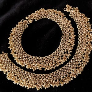 May include: Gold-toned anklets with intricate detailing. The anklets feature a repeating pattern of small, round embellishments and dangling pearl-like beads. The jewelry is displayed against a dark background.