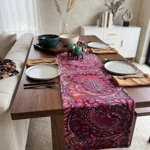 May include: A wooden dining table set with a paisley patterned table runner in shades of red, pink, and orange. The table is set with white plates, napkins, and silverware. Decorative teal elephant figurines and bowls are also present.