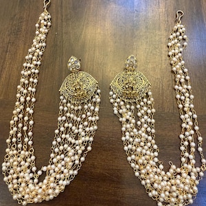 May include: A pair of gold-toned earrings with a domed, ornate design and dangling strands of small, white pearls. The earrings have a hook at the top and are set against a dark wooden surface.