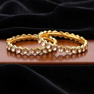 May include: Two gold-colored bangles with a geometric design. Each bangle features a series of clear, hexagonal-shaped stones set in a gold frame. The bangles are displayed on a wooden surface against a black backdrop.
