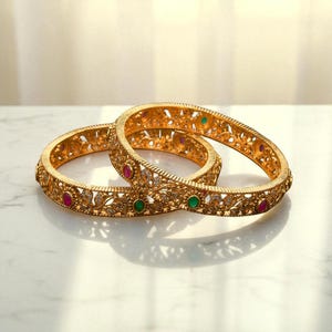 May include: Two gold bangles with intricate floral patterns and embedded gemstones. The bangles feature red, green, and clear gemstones. The bangles are displayed on a white surface.