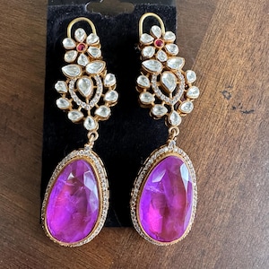 May include: A pair of gold dangle earrings with a large purple gemstone and a floral design with white and red stones.