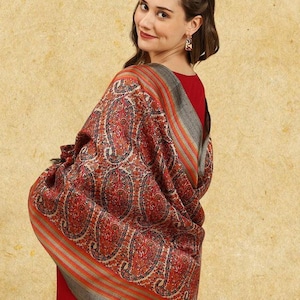 May include: A woman wearing a red dress and a shawl. The shawl features a paisley pattern in shades of red, orange, and gray. The shawl is draped over the shoulders and arms, creating a warm and stylish look. The background is a solid, neutral color.