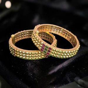 May include: Two gold-toned metal bangles featuring a textured pyramid stud pattern across the outer surface. Each bangle is accented with a small cluster of embedded round red and green stones and includes a hinged clasp mechanism for closure.
