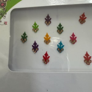 May include: A collection of colorful bindi forehead stickers in a white package. The stickers are leaf-shaped with a central jewel and come in various colors, including green, purple, yellow, red, and blue. The package has the word "DIAM" printed on it.