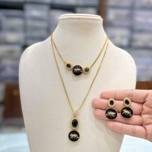 May include: Gold-toned jewelry set featuring a necklace and earrings. The necklace has a layered design with black circular and oval pendants, each displaying a gold animal silhouette. The earrings mirror the circular pendant design. The set is displayed on a mannequin.