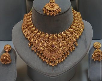 May include: A gold-toned jewelry set consisting of a wide, ornate necklace with paisley motifs and a central circular medallion, a matching maang tikka, and two bell-shaped jhumka earrings, all featuring intricate metalwork and small dangling beads.