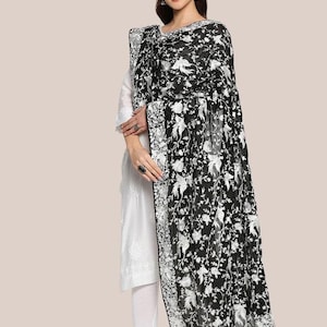May include: A black and white embroidered shawl with a floral pattern. The shawl is draped over a white outfit. The intricate design features white flowers and leaves against a black background, with a detailed border.