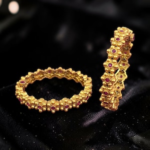 May include: Two circular gold bangles featuring a textured, beaded surface pattern with small, round, deep pink gemstone inlays. The bangles have a matte, antique-style finish and a repeating floral-inspired motif along the entire outer circumference.
