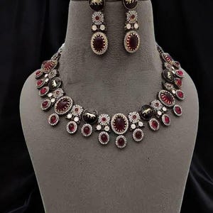 May include: A necklace and earring set featuring a dark metal finish. The necklace and earrings have oval and round red gemstones, surrounded by smaller white stones. The earrings and necklace also have black accents with a gold animal design.