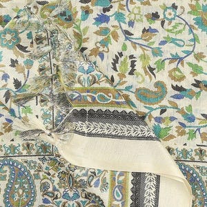 May include: Cream-colored textile with a detailed floral and paisley pattern in shades of blue, green, and brown. The fabric has a border with black and white geometric designs and tassels. The overall design is intricate and decorative.