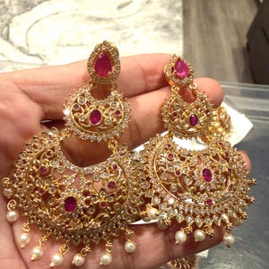 May include: Gold-toned chandelier earrings with intricate filigree and sparkling clear and ruby-colored stones. Small white pearls dangle from the bottom of the earrings. The earrings have a three-tiered design.