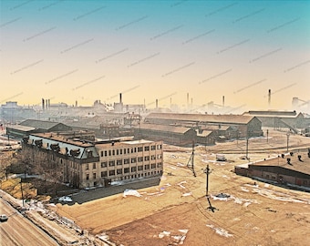 May include: An aerial view of a large industrial complex with a brick building in the foreground. The building has many windows and a flat roof. The complex is surrounded by a large, empty lot with a few trees and some snow on the ground.