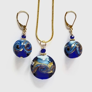 May include: A jewelry set consisting of a necklace and two matching earrings. Each piece features a round, deep blue glass bead with swirling patterns of light blue and gold metallic flecks. The necklace hangs on a thin gold-tone chain.