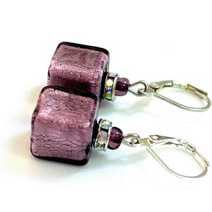 Pu&ograve; includere: Un paio di orecchini pendenti in argento con un ciondolo quadrato in vetro viola metallizzato e una perla di cristallo viola su ogni orecchino.