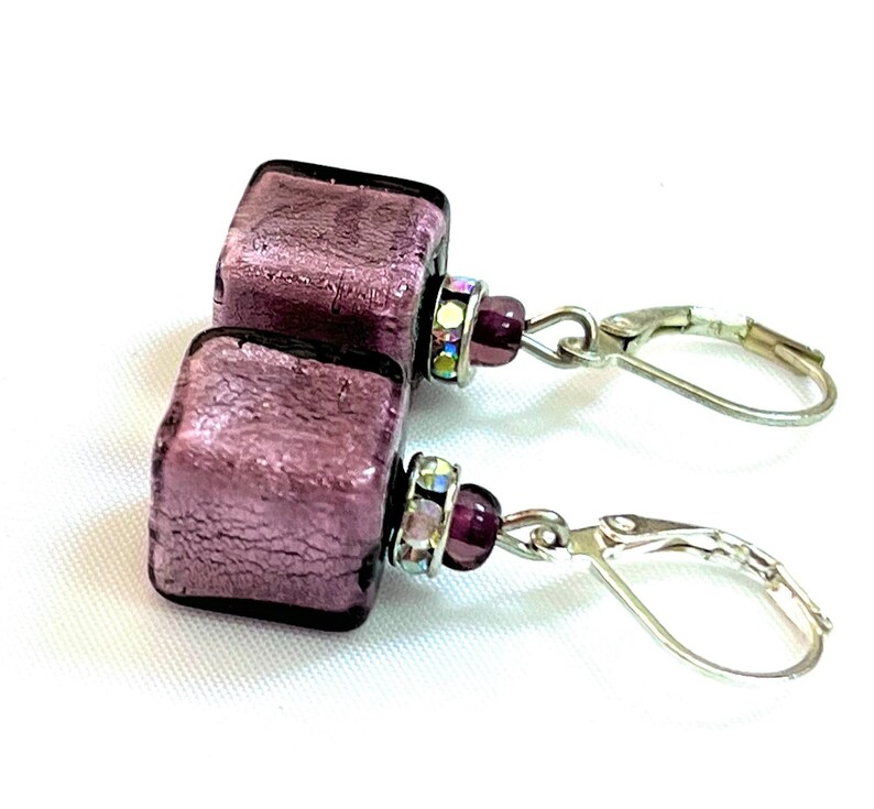 Pu&ograve; includere: Un paio di orecchini pendenti in argento con un ciondolo quadrato in vetro viola metallizzato e una perla di cristallo viola su ogni orecchino.