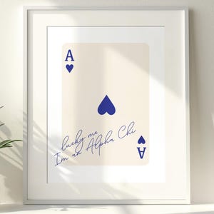 Può includere: Stampa incorniciata con un design di carte da gioco. La carta color crema mostra un simbolo di Asso di Picche blu e il testo "lucky me I'm an Alpha Chi" in blu. La stampa è in una cornice bianca.