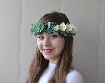 May include: A woman with long brown hair wearing a white shirt and a floral crown made of white flowers and green leaves.