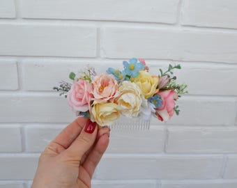 May include: A hair comb adorned with an arrangement of artificial flowers in shades of pastel pink, cream, yellow, and light blue, accented with small green leaves and white filler flowers, featuring a clear plastic comb base for hair attachment.