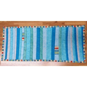 May include: A rectangular, handmade quilt with vertical stripes in shades of blue and grey. The quilt has a patterned border with red, green, and blue accents. Two small, decorative patches with geometric patterns are centered on the quilt.