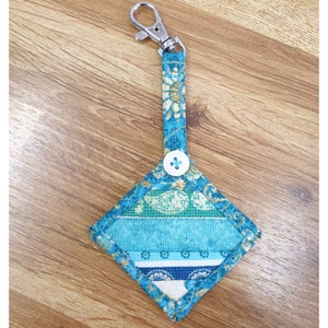 Op de afbeelding: Een teal blauwe en witte stoffen sleutelhanger met een ruitvormige hanger en een knoopsluiting. De hanger is gemaakt van patchwork stof met een bloemenpatroon.