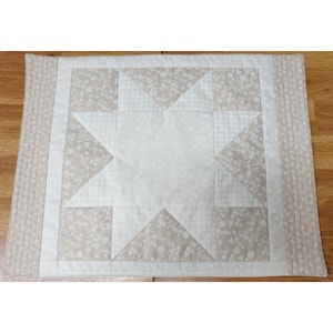 May include: A beige and white quilted table runner with a star design. The star is made up of smaller squares and triangles.
