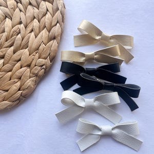 May include: Five leather hair bows in different colors. Two gold bows, one black bow, and two white bows.