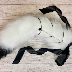 May include: A fluffy white and black faux fur tail with a black satin ribbon tie. The tail is accented with a silver chain detail. The tail is a costume accessory.