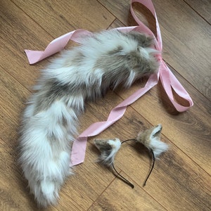 Op de afbeelding: Een faux fur staart en bijpassende hoofdband met oren. De staart is wit, grijs en bruin en is vastgebonden met een roze satijnen lint. De hoofdband heeft kleine oren met hetzelfde vachtpatroon.