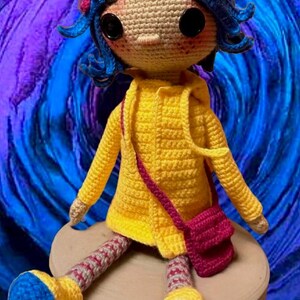 May include: A crocheted doll with blue hair, wearing a yellow raincoat and a pink bag, sitting on a round platform against a swirling purple and blue background.