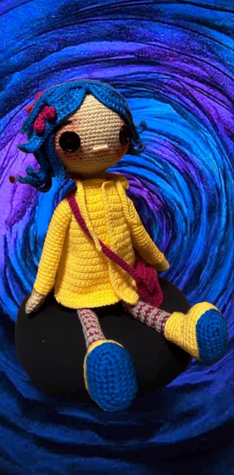 May include: A crocheted doll with blue hair, a yellow jacket, and pink and white striped pants. The doll is sitting on a black surface with a blue and purple swirl background.