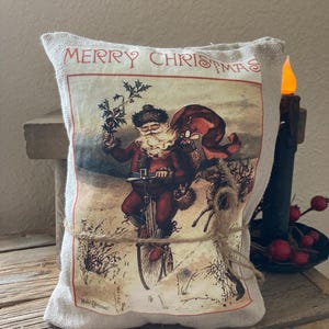 May include: A rectangular, beige Christmas pillow with a vintage illustration of Santa Claus riding a bicycle, carrying a sack. The words "MERRY CHRISTMAS" are printed at the top. The pillow is tied with twine and sits on a wooden surface.