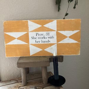 May include: A rectangular wooden sign with a yellow and white geometric pattern. The text "Prov. 31 She works with her hands" is centered on the sign. A small wooden stool and a black candle holder with a black candle are in the foreground.