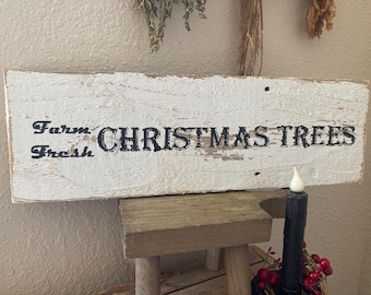 May include: A rustic, rectangular wooden sign with the words "Farm Fresh CHRISTMAS TREES" in black lettering. The sign is painted white with a distressed finish. A black candle with red berries sits below the sign.
