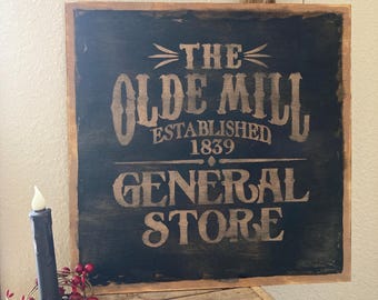 May include: A rustic wooden sign with the text "THE OLDE MILL ESTABLISHED 1839 GENERAL STORE" in a distressed font. A black candle with a red berry wreath sits to the left. The sign is on a wooden surface.
