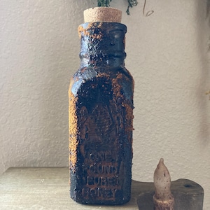 May include: A dark glass bottle with a cork stopper and a rusty brown patina. The bottle has a label that reads "One Pound Pure Honey".