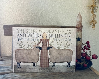 Peut inclure: Une pancarte en bois rustique avec un fond beige et des lettres brunes. La pancarte représente une femme en robe à carreaux rouge et noir debout entre deux moutons. Le texte sur la pancarte dit "She seeks wool and flax and works willingly with her hands Proverbs 31:13".