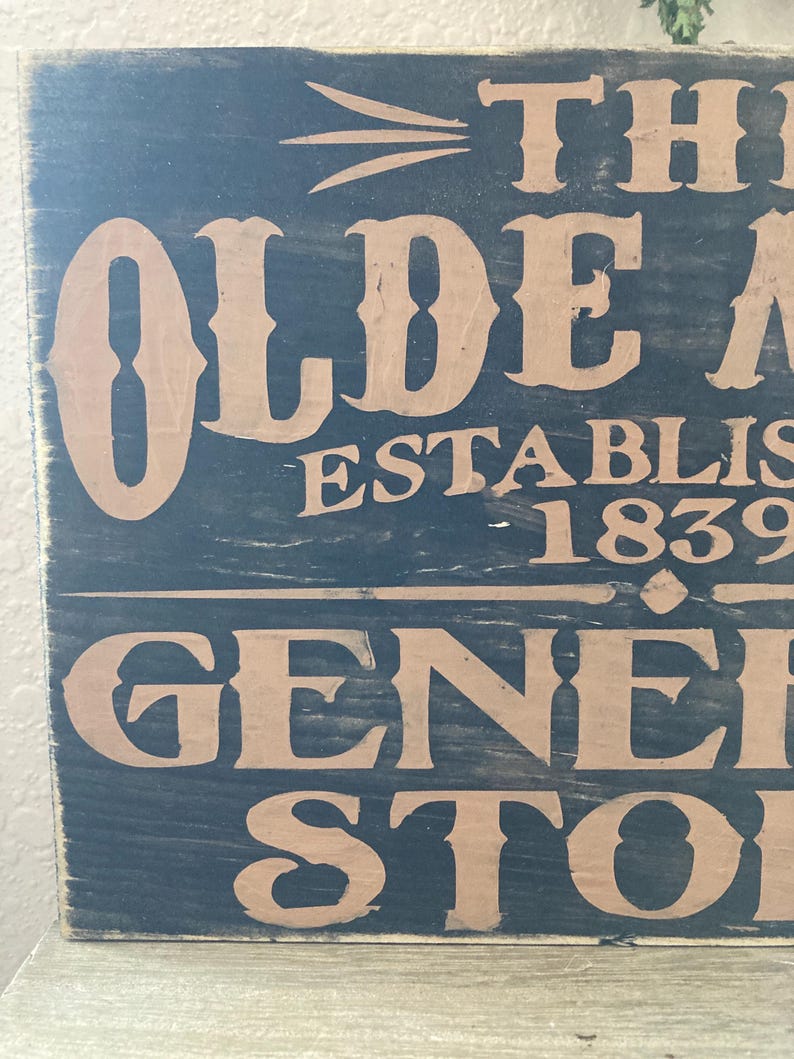 May include: A black wooden sign with the text "The Olde Mill Established 1839 General Store" in brown lettering.