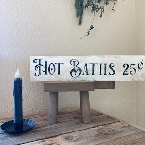 May include: A rustic wooden sign with the words "HOT BATHS 25¢" in black lettering. The sign rests on a small wooden stool. A dark blue candle sits in a black metal holder to the left of the sign. Dried greenery hangs above.